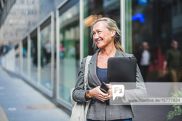 Happy mature businesswoman looking away while standing against office building