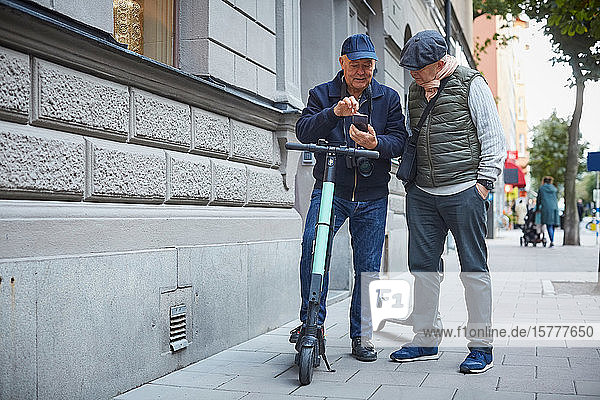 Homosexuelles Paar in voller Länge  das ein Mobiltelefon benutzt  während es mit Rollern auf dem Bürgersteig in der Stadt steht