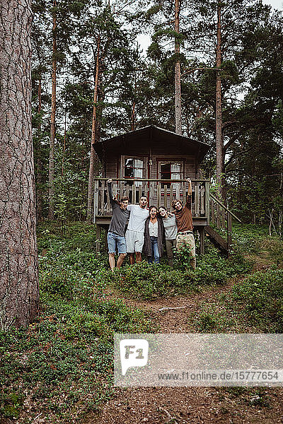 Fröhliche Freunde  die während des Urlaubs mit den Armen um das Haus im Wald stehen