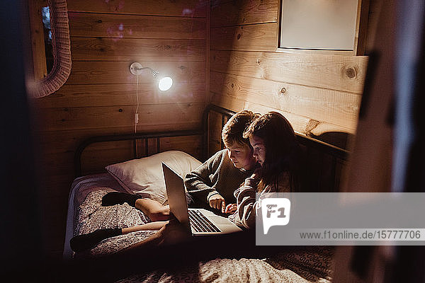 Bruder und Schwester benutzen einen Laptop  während sie zu Hause im Schlafzimmer an der Wand sitzen