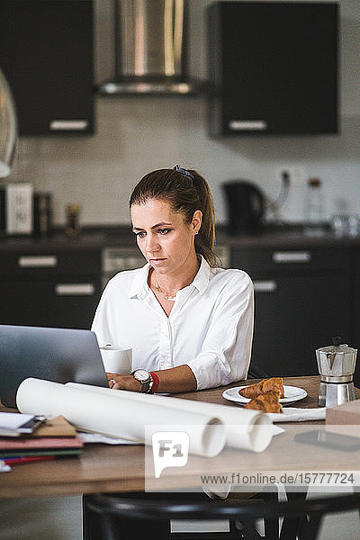 Weibliche Freiberuflerin benutzt Laptop  während sie zu Hause mit Kaffeetasse sitzt