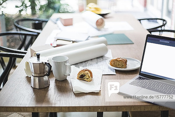 Hochwinkelansicht einer Teekanne mit aufgerollten Dokumenten per Laptop und Croissant auf dem Tisch