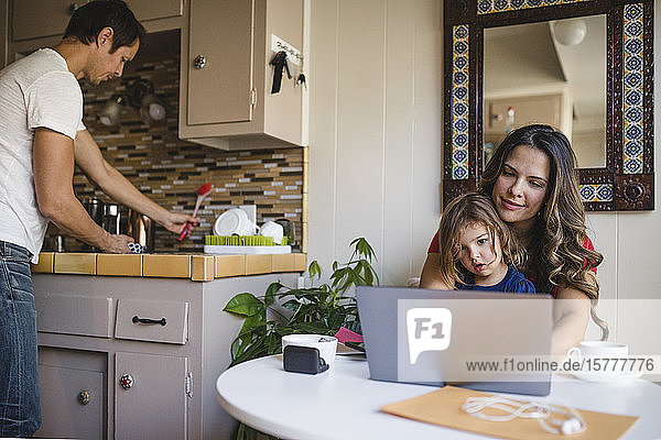 Mutter und Tochter benutzen Laptop  während der Vater in der Küche arbeitet