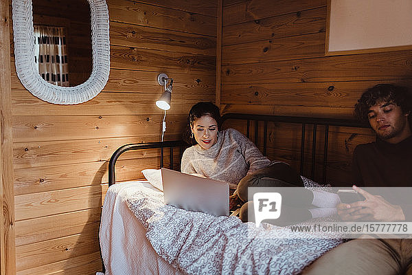 Junge Frau benutzt Laptop  während sie mit einem Freund zu Hause im Bett sitzt