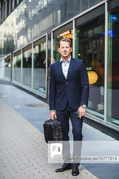 Portrait of mature businessman with bag standing against office building