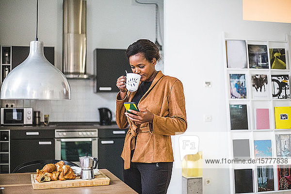 Smiling female architect drinking while using phone at home