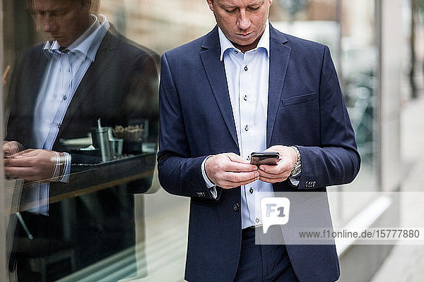 Kontemplation eines reifen Geschäftsmannes  der ein Smartphone benutzt  während er am Glasfenster steht