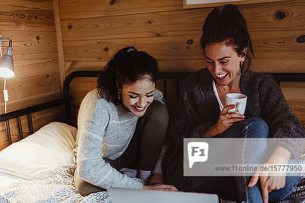 Smiling young woman using laptop while sitting with friend on bed at home