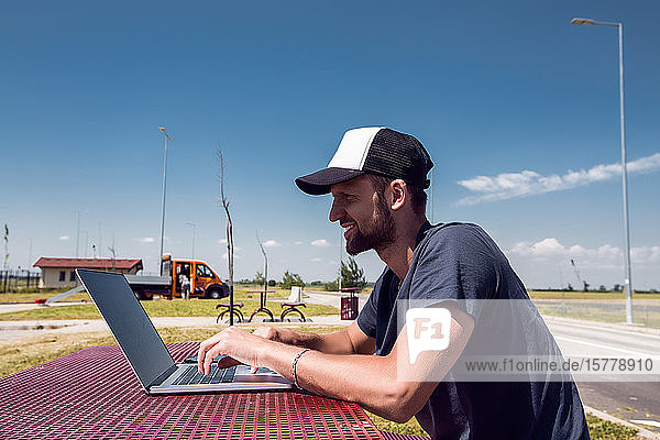 Mann mit Baseballmütze sitzt am Picknicktisch am Straßenrand und tippt am Laptop