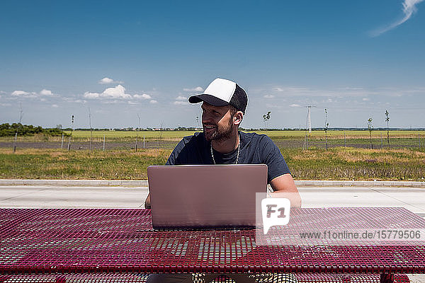 Mann mit Baseballmütze sitzt mit Laptop am Picknicktisch am Straßenrand