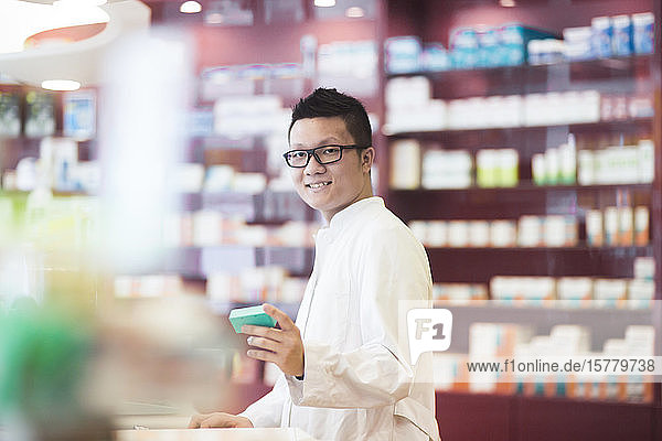 Male Asian pharmacist wearing glasses standing in front of shelves with medicines in a pharmacy.