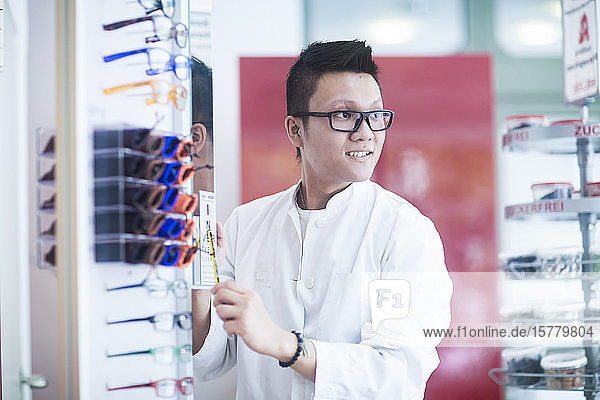 Männlicher asiatischer Apotheker mit Brille  der in der Apotheke neben einer Auslage mit Lesebrillen steht.