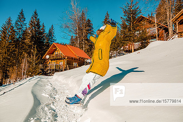 Junge Frau springt rückwärts in den tiefen Schnee in der Nähe einer Hütte.