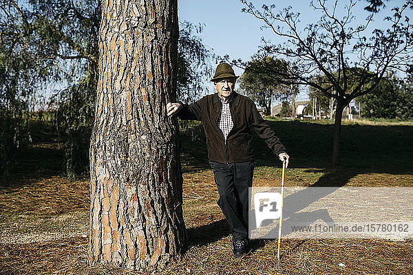 Old man with cane leaning on tree in park