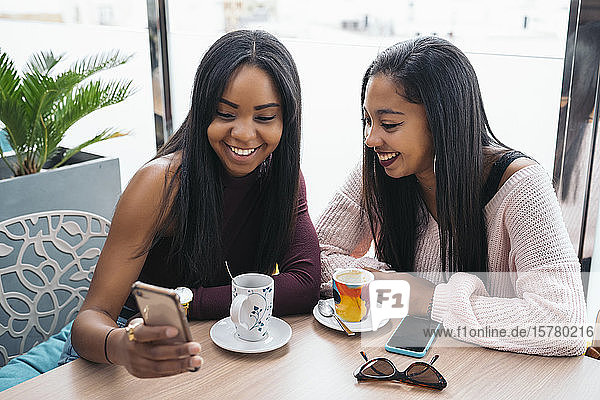 Two happy young women sharing cell phone in a cafe
