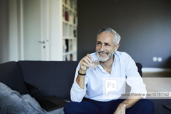 Lächelnder reifer Mann  der zu Hause auf dem Sofa ein Glas Wasser trinkt
