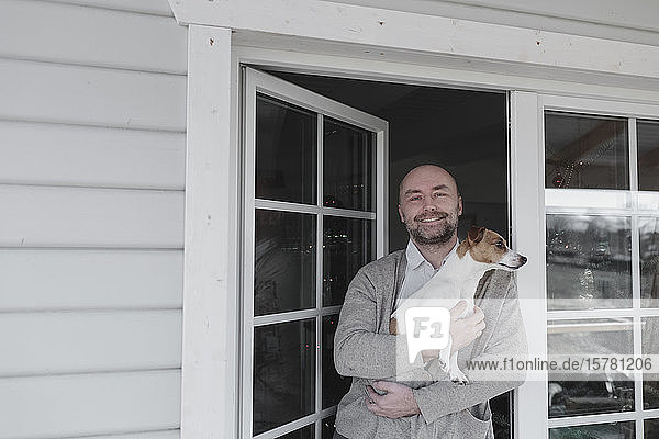 Porträt eines glücklichen Mannes mit Hund auf den Armen  der an der offenen Terrassentür seines Hauses steht