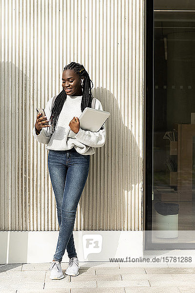 Junge Frau steht mit Handy und Laptop an einer Wand