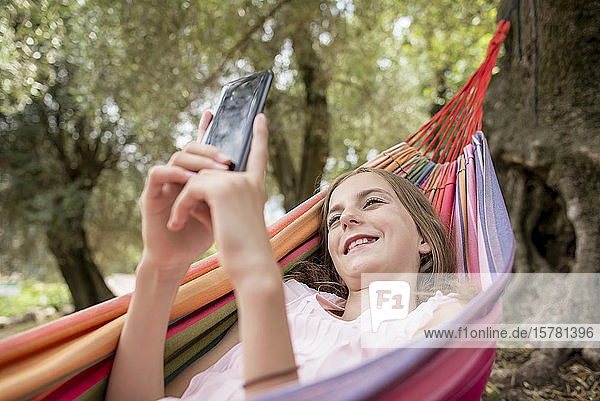 Mädchen liegt in Hängematte in einem Olivenhain und benutzt ein Smartphone