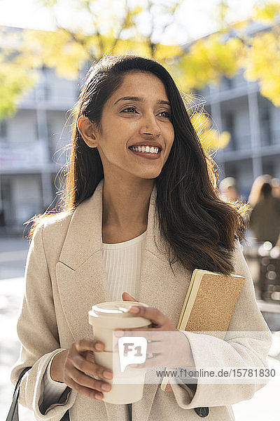 Lächelnde junge Frau mit Buch und Kaffee zum Mitnehmen in der Stadt  die sich umsieht