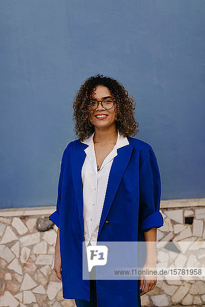 Porträt einer lächelnden Frau mit Brille und blauem Blazer  blaue Wand im Hintergrund