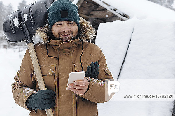 Porträt eines lächelnden Mannes mit Schneeschaufel  der auf sein Handy schaut