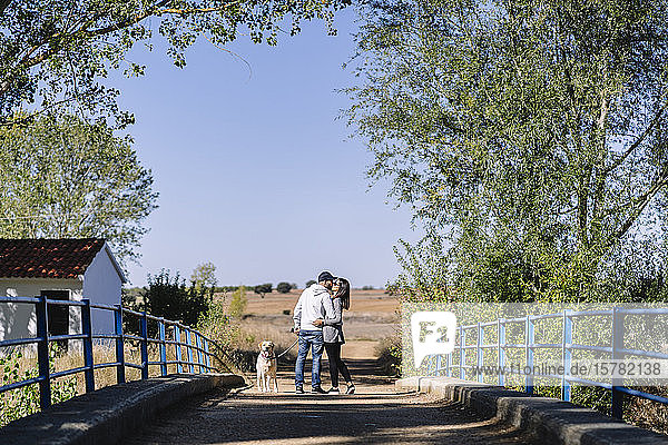 Ein küssendes Paar mit seinem Hund  das auf einer Brücke in einem Park steht
