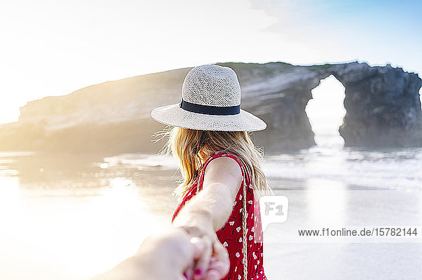 Blond woman wearing red dress and hat and holding hand on the beach  Natural Arch at Playa de Las Catedrales  Spain