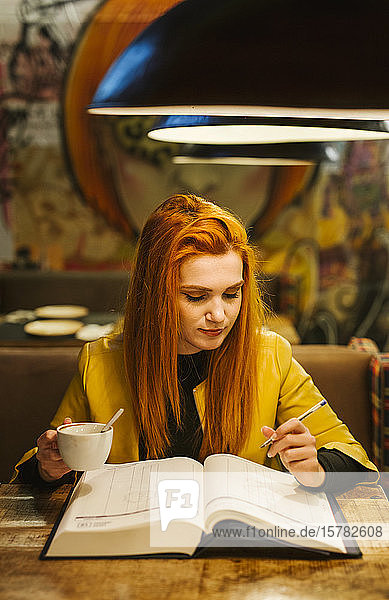 Porträt einer rothaarigen jungen Frau mit Buch beim Kaffeetrinken in einer Kneipe
