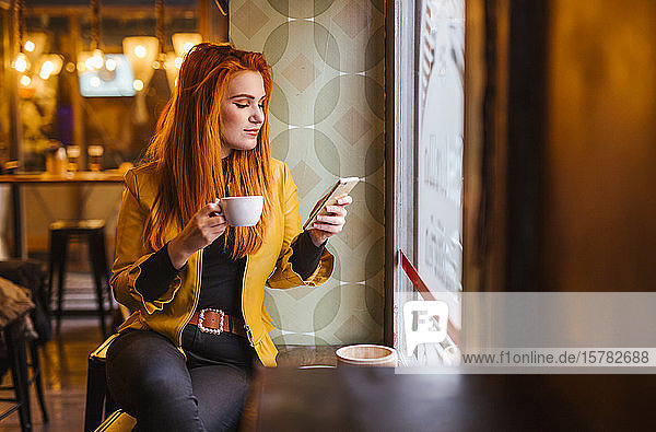 Rothaarige junge Frau sitzt in einem Cafe mit einer Tasse Kaffee und schaut auf ihr Handy