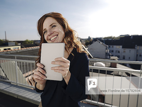 Glückliche rothaarige Frau mit Tablette auf der Dachterrasse