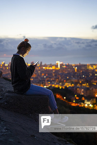 Junge Frau sitzt auf einem Geländer über der Stadt und telefoniert per Handy  Barcelona  Spanien