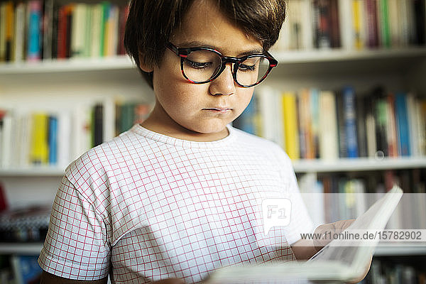 Porträt eines ernsthaften Jungen mit Brille  der vor einem Bücherregal steht und einen Comic liest