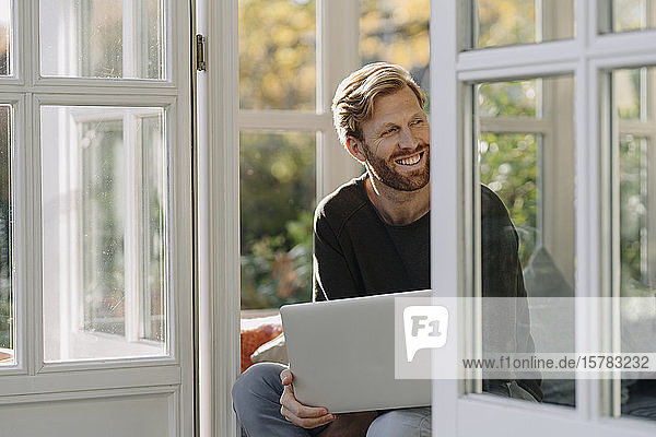 Lächelnder Mann mit Laptop im Wintergarten zu Hause