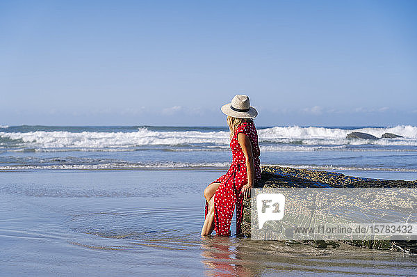 Blonde Frau in rotem Kleid und Hut sitzt auf einem Felsen am Strand Playa de Las Catedrales Spanien