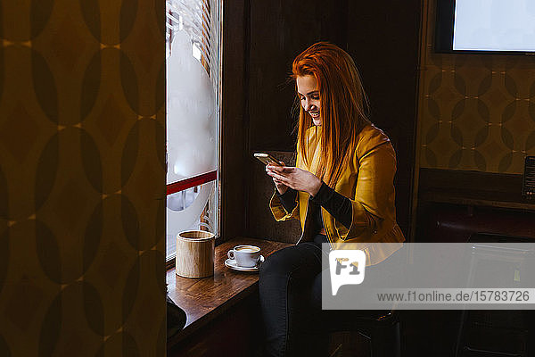 Glückliche rothaarige junge Frau sitzt in einem Café und benutzt ihr Handy
