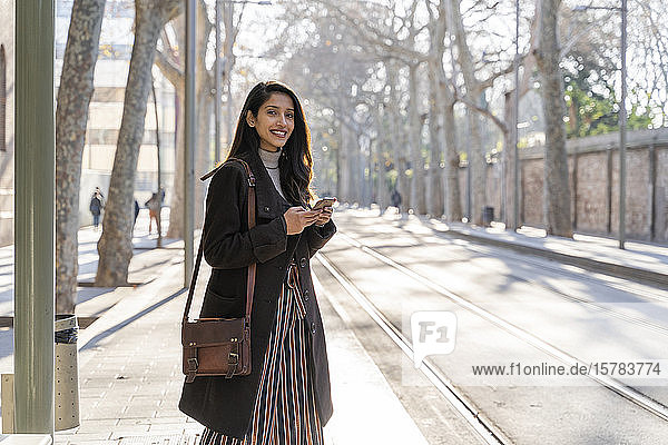 Porträt einer lächelnden jungen Frau mit Smartphone an der Straßenbahnhaltestelle