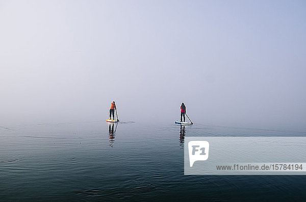 Zwei Frauen stehen beim Paddelsurfen auf einem See im Nebel auf
