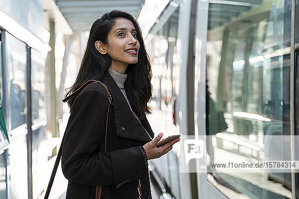 Lächelnde junge Frau mit Smartphone an der Straßenbahnhaltestelle