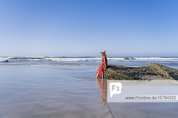 Blonde Frau in rotem Kleid und Hut sitzt auf einem Felsen am Strand  Playa de Las Catedrales  Spanien