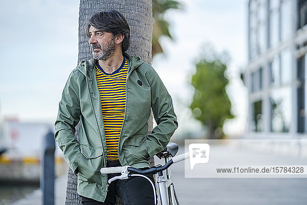 Porträt eines seriösen reifen Mannes mit einem Fahrrad  das an einen Palmenstamm gelehnt ist und in die Ferne schaut  Alicante  Spanien