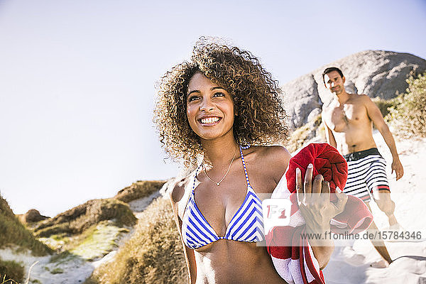 Porträt einer lächelnden jungen Frau auf dem Weg zum Strand mit einem Mann im Hintergrund