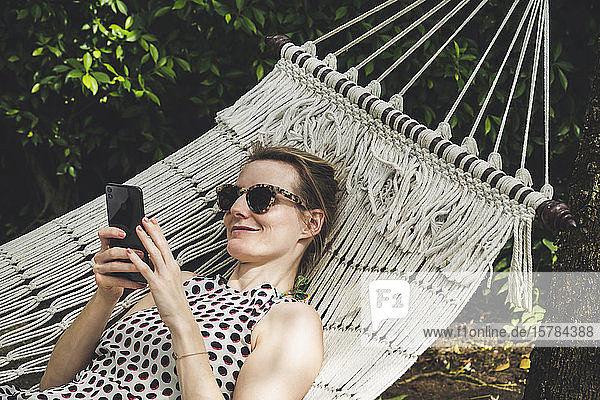 Lächelnde Frau liegt in einer Hängematte und benutzt Mobiltelefon