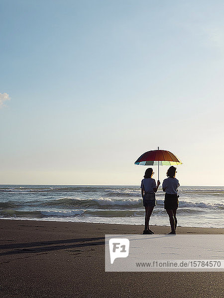 Zwei Frauen mit Sonnenschirm stehen in der Abenddämmerung am Strand  Kedungu Beach  Bali  Indonesien