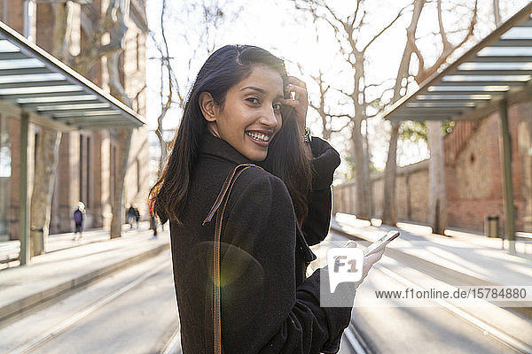 Porträt einer glücklichen jungen Frau mit Smartphone auf der Straßenbahnlinie