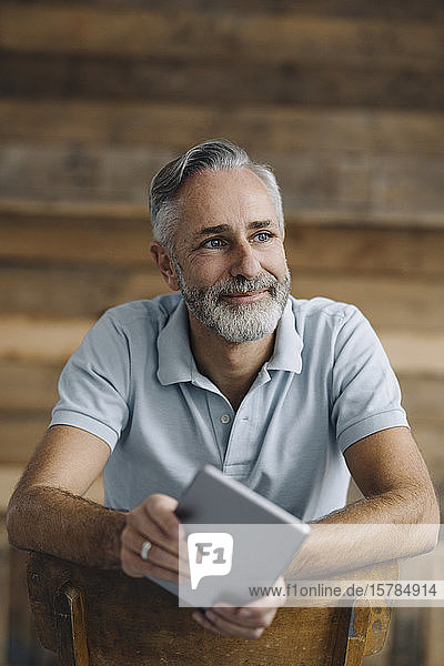 Porträt eines lächelnden reifen Mannes mit digitalem Tablett  der sich auf die Rückenlehne lehnt und in die Ferne schaut