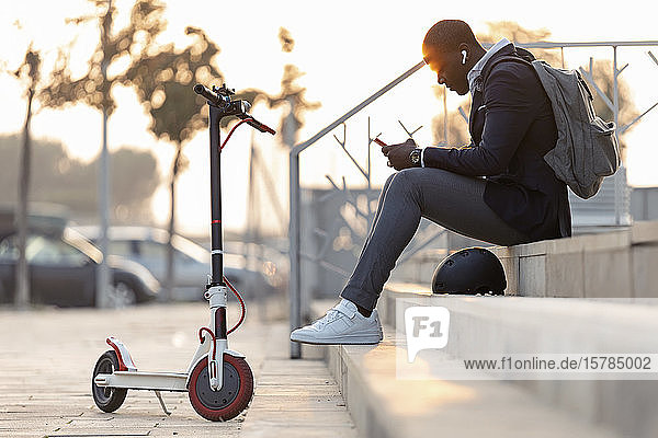 Junger Geschäftsmann mit Schubroller  der bei Sonnenuntergang mit Mobiltelefon und Kopfhörern auf der Treppe sitzt