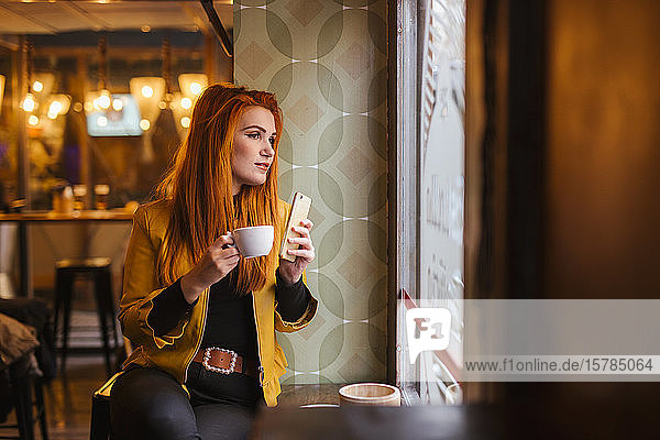 Porträt einer rothaarigen jungen Frau mit Smartphone  die in einem Café wartet und aus dem Fenster schaut