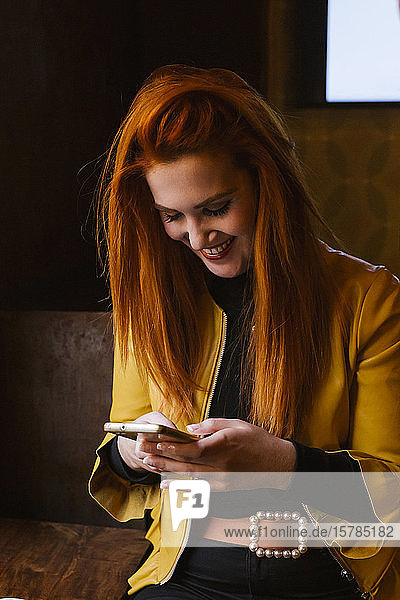 Porträt einer glücklichen rothaarigen jungen Frau in einer Kneipe  die auf ihr Handy schaut