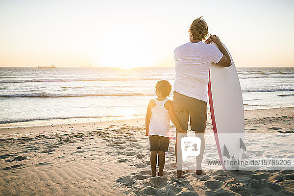 Rückansicht von Vater und Sohn mit Surfbrett  die bei Sonnenuntergang am Strand stehen